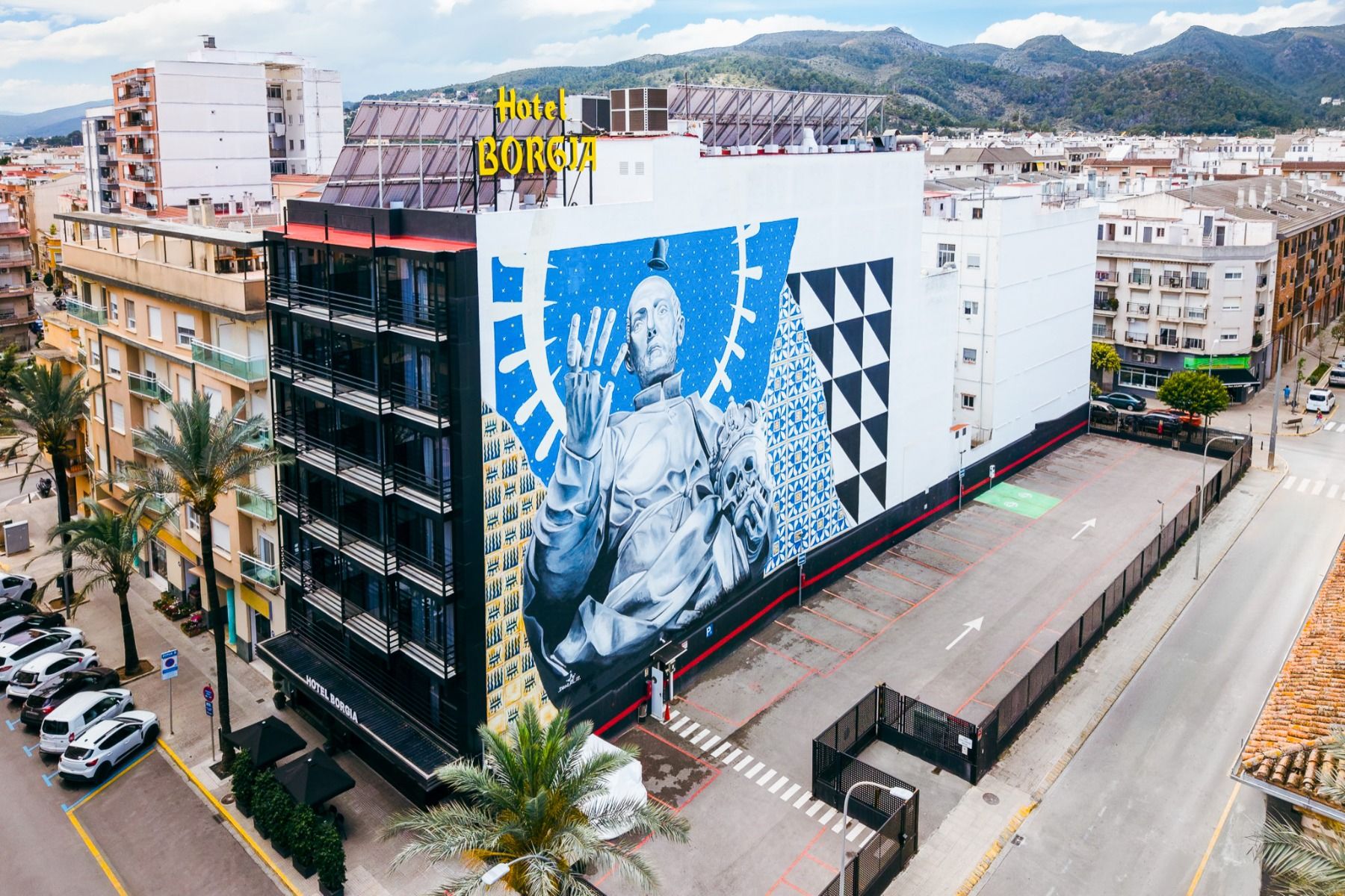 A large mural of a futuristic figure adorns the facade of the Hotel Borgia *, which overlooks a largely empty parking lot, set amid an urban landscape framed by hills and buildings. The Hotel Borgia *, known for hosting soccer teams during their training