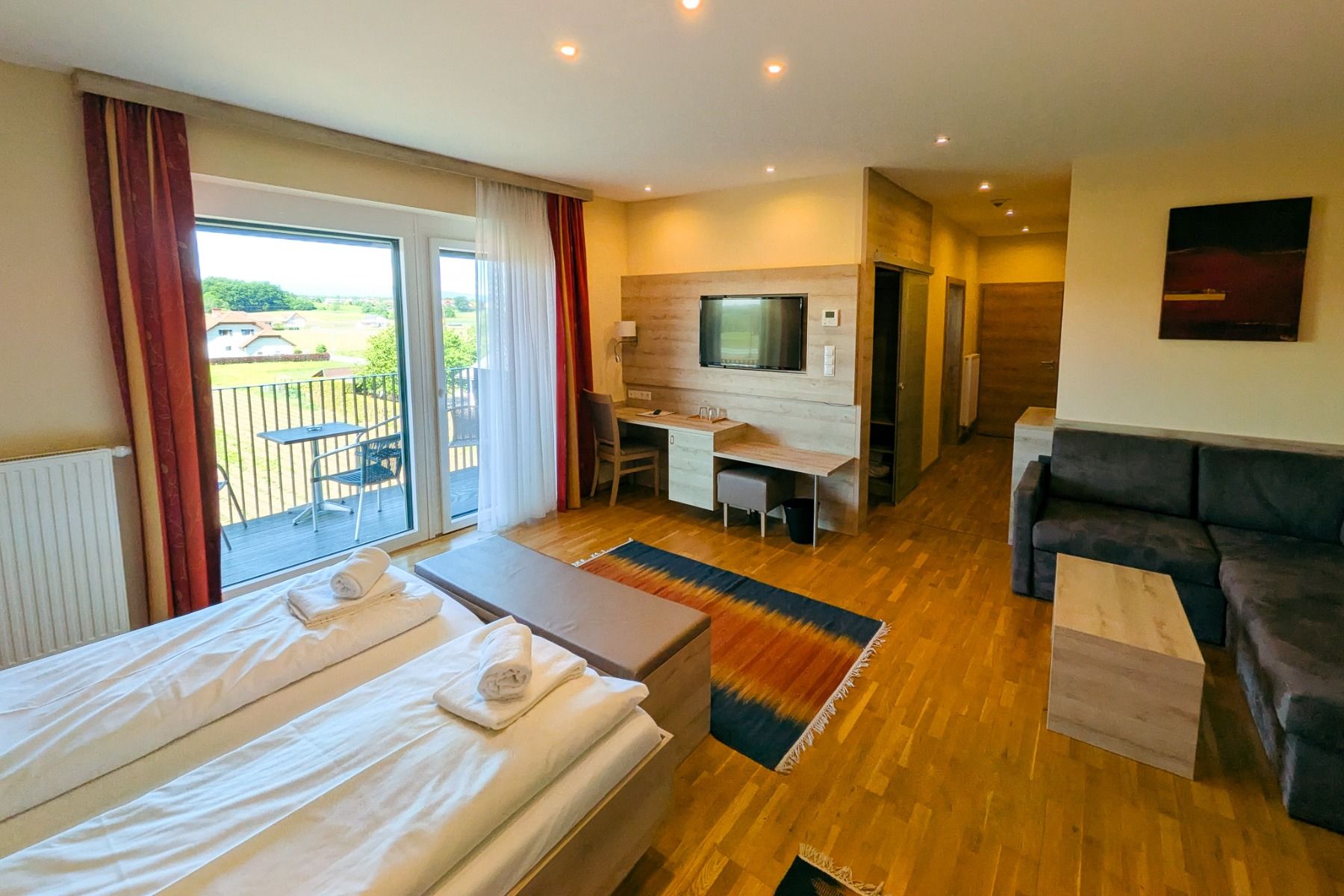 The Hotel Restaurant Schachenwald offers a modern room with hardwood floors, a double bed, a sofa, a desk, a TV, and patio doors overlooking the countryside—ideal for guests at the training camp or visitors to Schwarzsee—featuring warm lighting and colorf