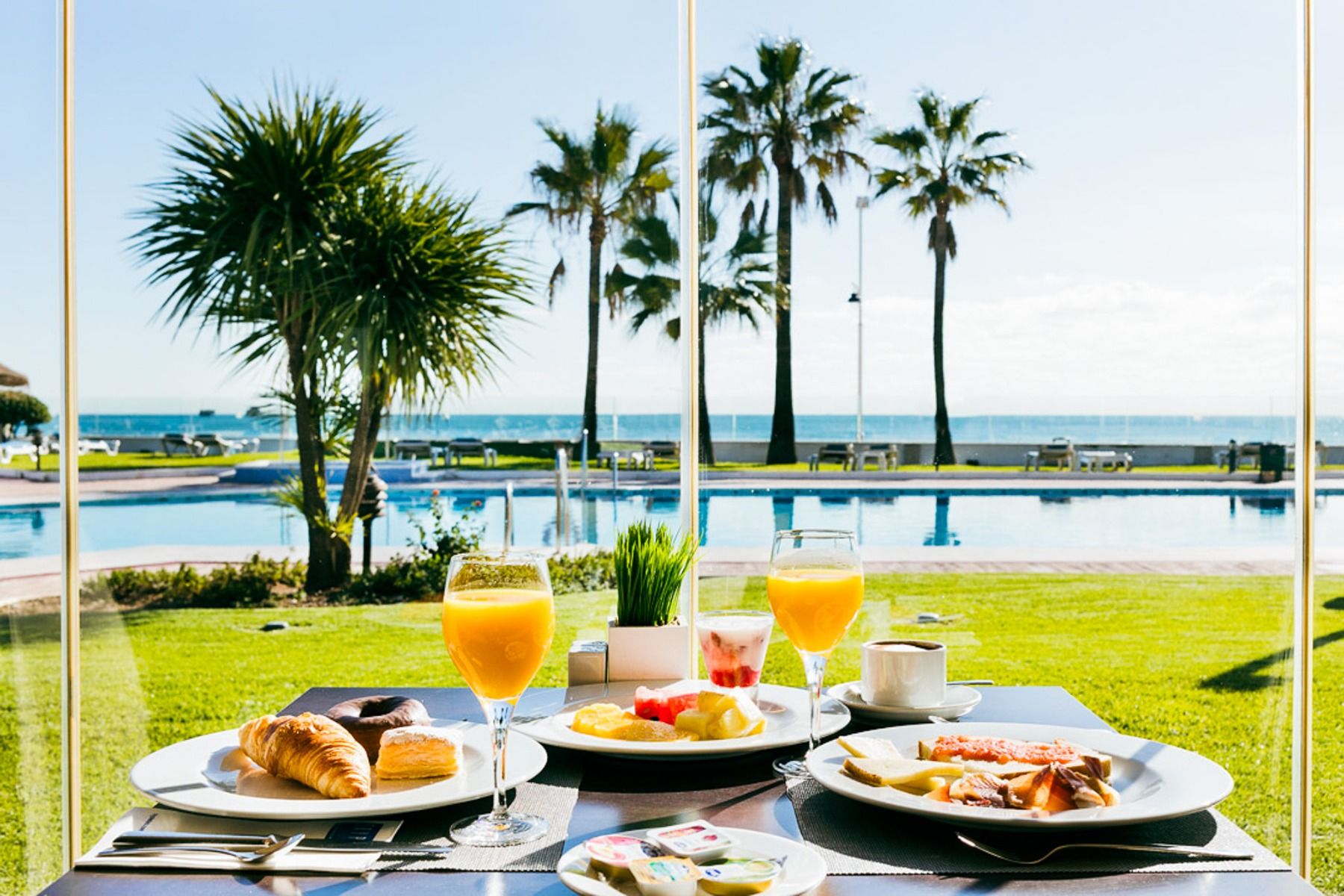 A breakfast table with croissants, fruit, juice and coffee at the Hotel Sol Guadalmar offers a view of the pool, the green lawn, the palm trees and the sea in the background on a sunny day.
