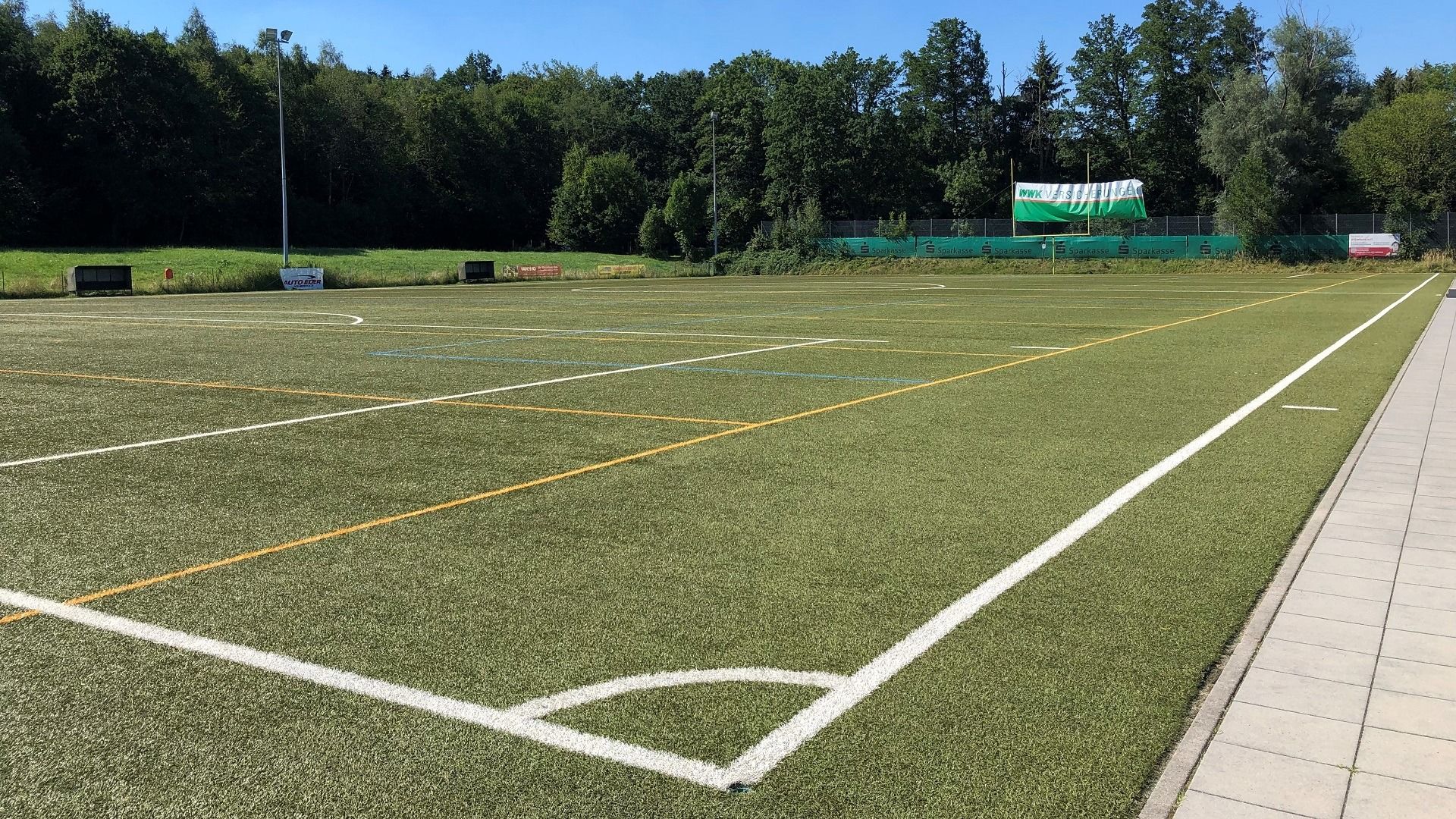 Sportbund DJK Rosenheim has an empty artificial turf pitch with white and yellow lines, surrounded by trees and green areas under a clear blue sky. A paved walkway runs along the right-hand side of the pitch.