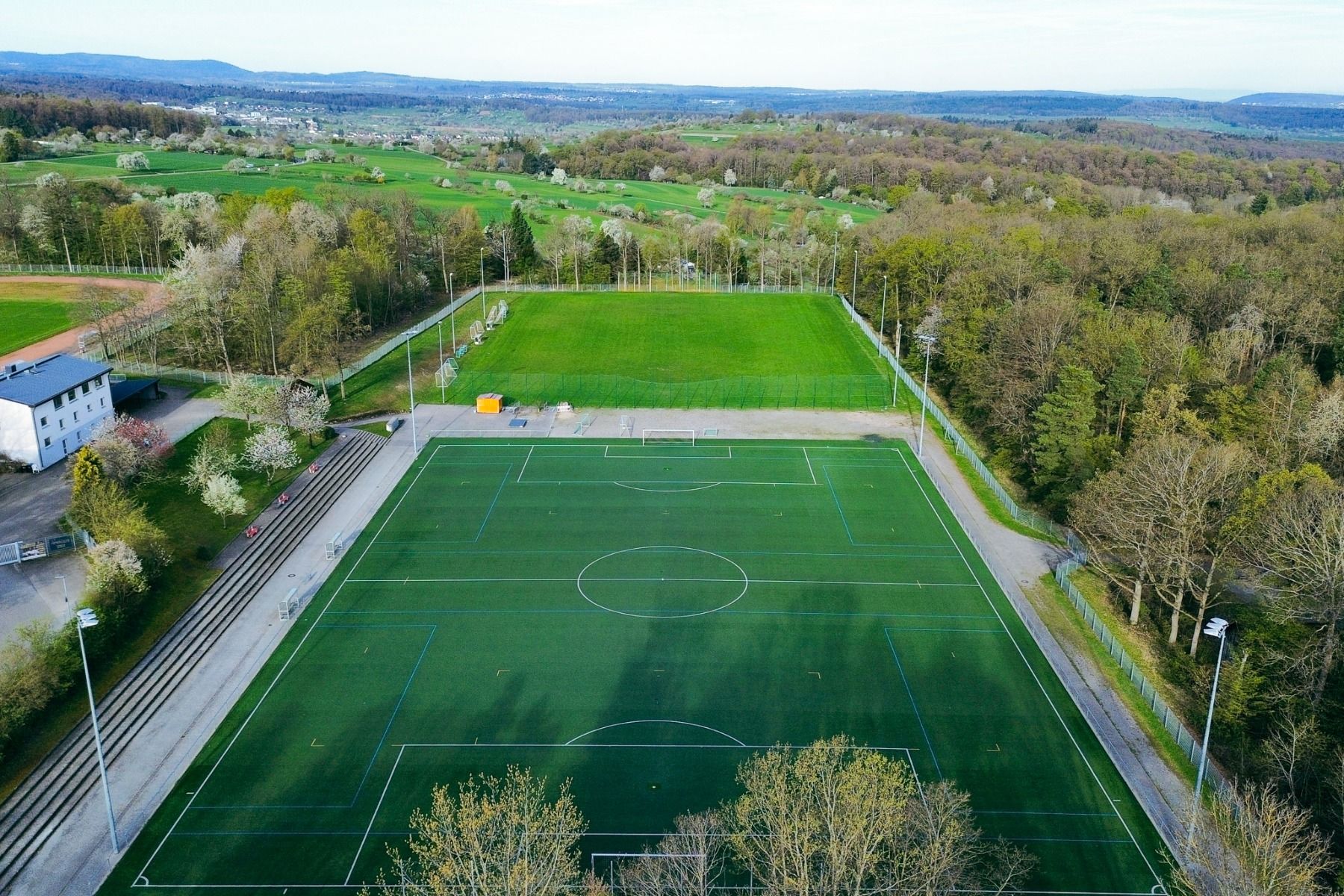 Aerial view of a green artificial turf field with white markings, surrounded by trees, with bleachers on one side and a building nearby—nestled in a rural landscape with hills in the distance— which offers teams looking for the ideal location for a traini