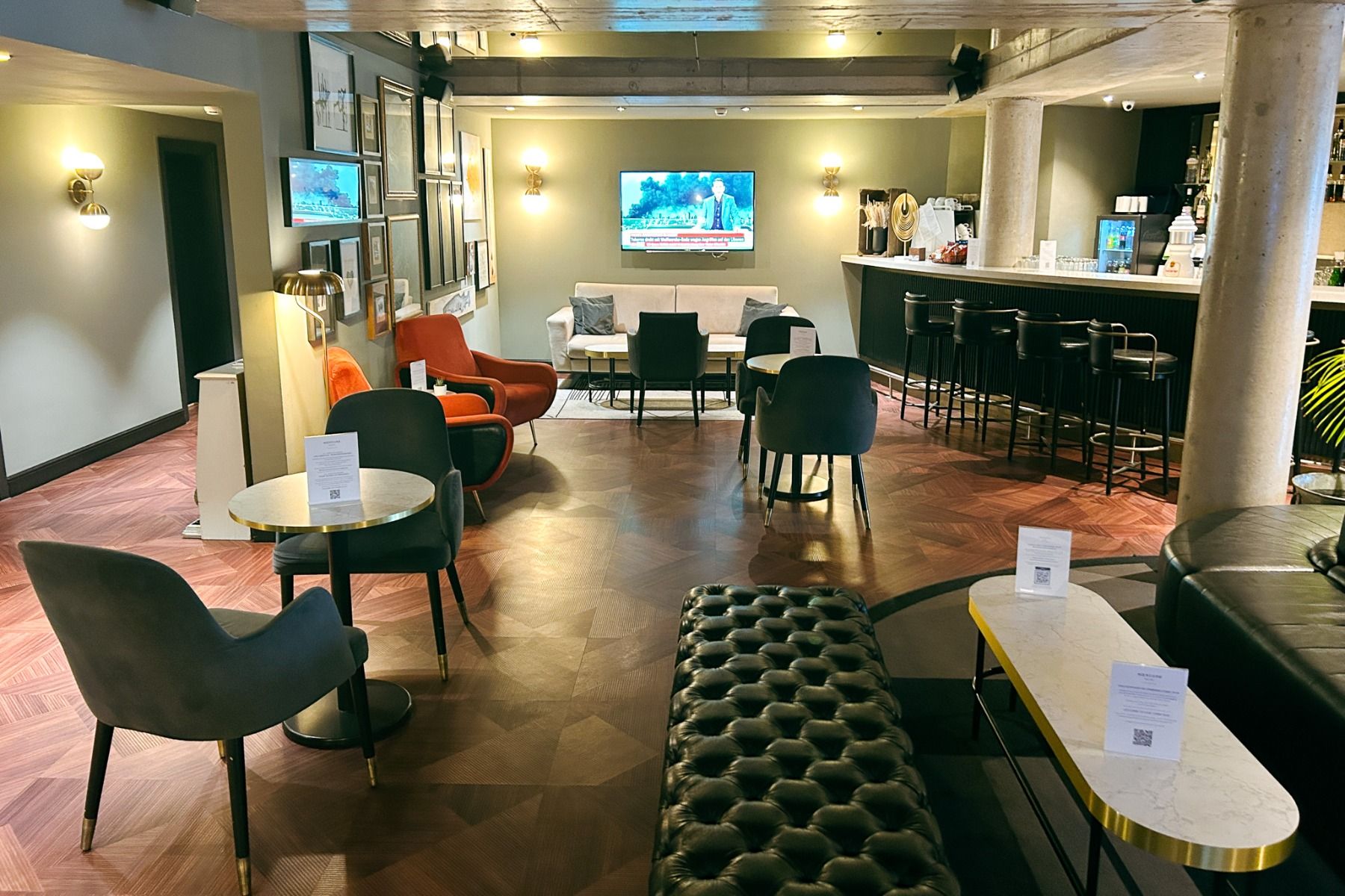 Enjoy the modern lounge at the Mercure Hotel Pforzheim, featuring a selection of comfortable chairs, sofas, stylish tables, a well-stocked bar with bar stools, colorful murals, and TV screens showing the latest news. Warm lighting and hardwood floors crea