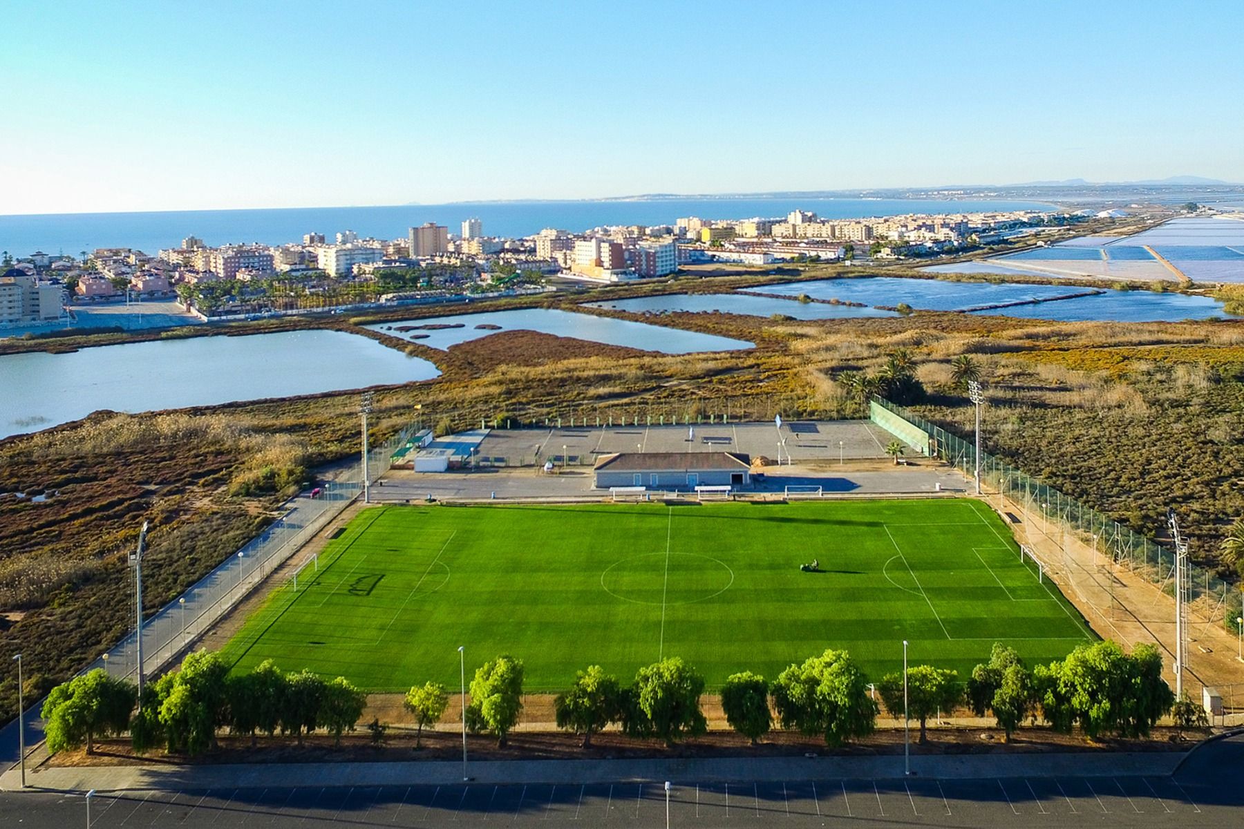 Aerial view of a green soccer pitch in Santa Pola, lined with trees, stands and buildings near wetlands and an urban landscape under a blue sky, near the Hotel Gaudea.