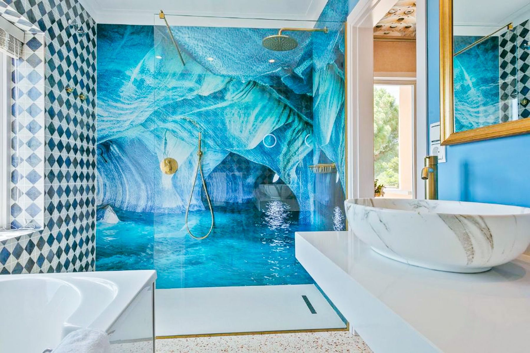 Modern bathroom in the Van der Valk Hotel BARCAROLA with blue walls, white marble washbasin and a shower with an ice-blue photo wallpaper of a cave. Golden fittings and geometric tiles are reminiscent of the luxury of the beautiful coasts of the Costa Bra