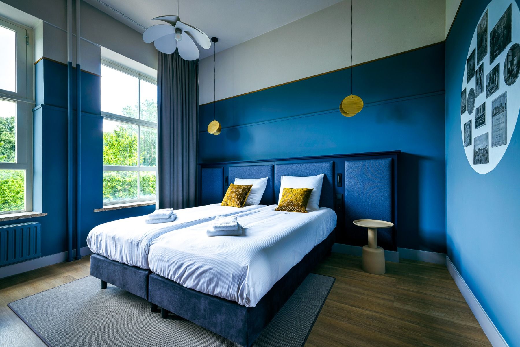 A modern room at Watersley Sports & Talentpark with two single beds, white bedding, mustard yellow pillows, blue walls, round lamps, large windows with gray curtains, wooden floor and a photo collage - very close to Sportcampus Sittard.