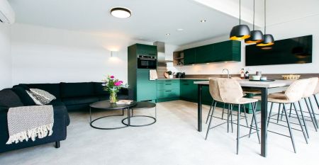 Duinrell: Modern two-story house with large windows, yellow and gray exterior, covered patio with seating. Located near top sports facilities, it features a green lawn and trees under a partly cloudy sky.