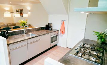 Duinrell: Modern two-story house with large windows, yellow and gray exterior, covered patio with seating. Located near top sports facilities, it features a green lawn and trees under a partly cloudy sky.