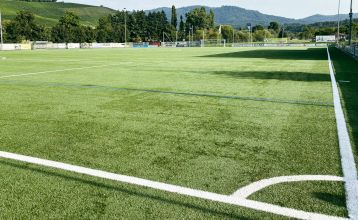 Aerial view of Hanauer Hof, a corner hotel with a brown tiled roof and solar panels, located near an artificial turf pitch in Appenweier, surrounded by cars and streets in a small town on a sunny day.