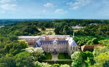 Aerial view of Watersley Sports & Talent Park, a sprawling historic gray-roofed complex nestled in a verdant landscape. The campus includes a chapel-like building, world-class training facilities and offers views of distant fields under clear skies.