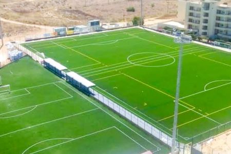Aerial view of “Campo David Neto N2” in Portimão, showing two adjacent soccer fields with artificial turf and white and yellow field markings, surrounded by a fence; buildings and dry land can be seen in the background.