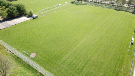 Aerial view of training pitch 1 Baunatal: an empty, well-kept soccer pitch with marked lines, surrounded by a fence and trees. Benches and small buildings can be seen at the edge of the pitch.
