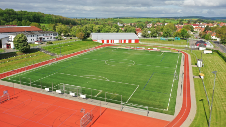 A bright yellow corner flag stands at the intersection of white lines on the Schlotheim artificial turf pitch, a large green artificial turf soccer pitch with buildings and trees in the background under a blue sky.