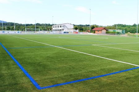 SC Durbachtal's artificial turf pitch 2 has a green artificial turf with white and blue markings, two buildings in the background and is surrounded by trees and fences in the open air.