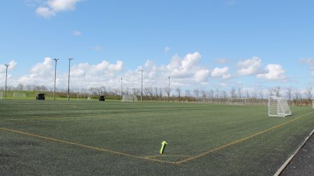 An empty outdoor soccer pitch with artificial turf from Kunstrasen Skaerbaek, several goalposts, high light poles under a blue sky with scattered clouds and a green cone near the foreground.