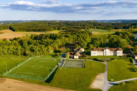 Aerial view of the Schielleiten artificial turf pitch with sports fields, tennis courts and a large complex of buildings, surrounded by trees and open fields under a partly cloudy sky.