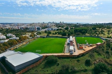 Aerial view of the natural-grass stadium “Estádio Municipal dos Dois Irmãos,” a lush green soccer complex surrounded by trees and buildings, with a cityscape and the sea in the background under a partly cloudy sky.