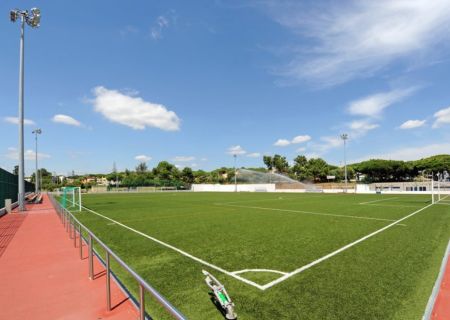Estadio Municipal Quarteria N2 shows an empty outdoor soccer field with artificial turf, bright white boundary lines, and a sprinkler system running in the background, while scattered clouds are visible against the blue sky. One side of the field is borde