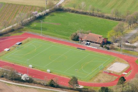 Aerial view of the “Ortenberg Artificial Turf Field” sports complex, featuring a soccer field with green artificial turf, surrounded by a red running track, bleachers, and a small building next to grassy areas and sports fields.