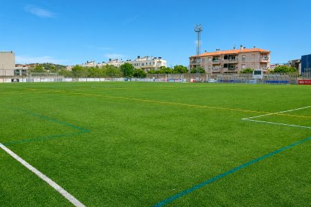 Wide view of the empty Camp Municipal de Futbol Vilartagues with green artificial turf, white and yellow lines and buildings in the background under a clear blue sky.