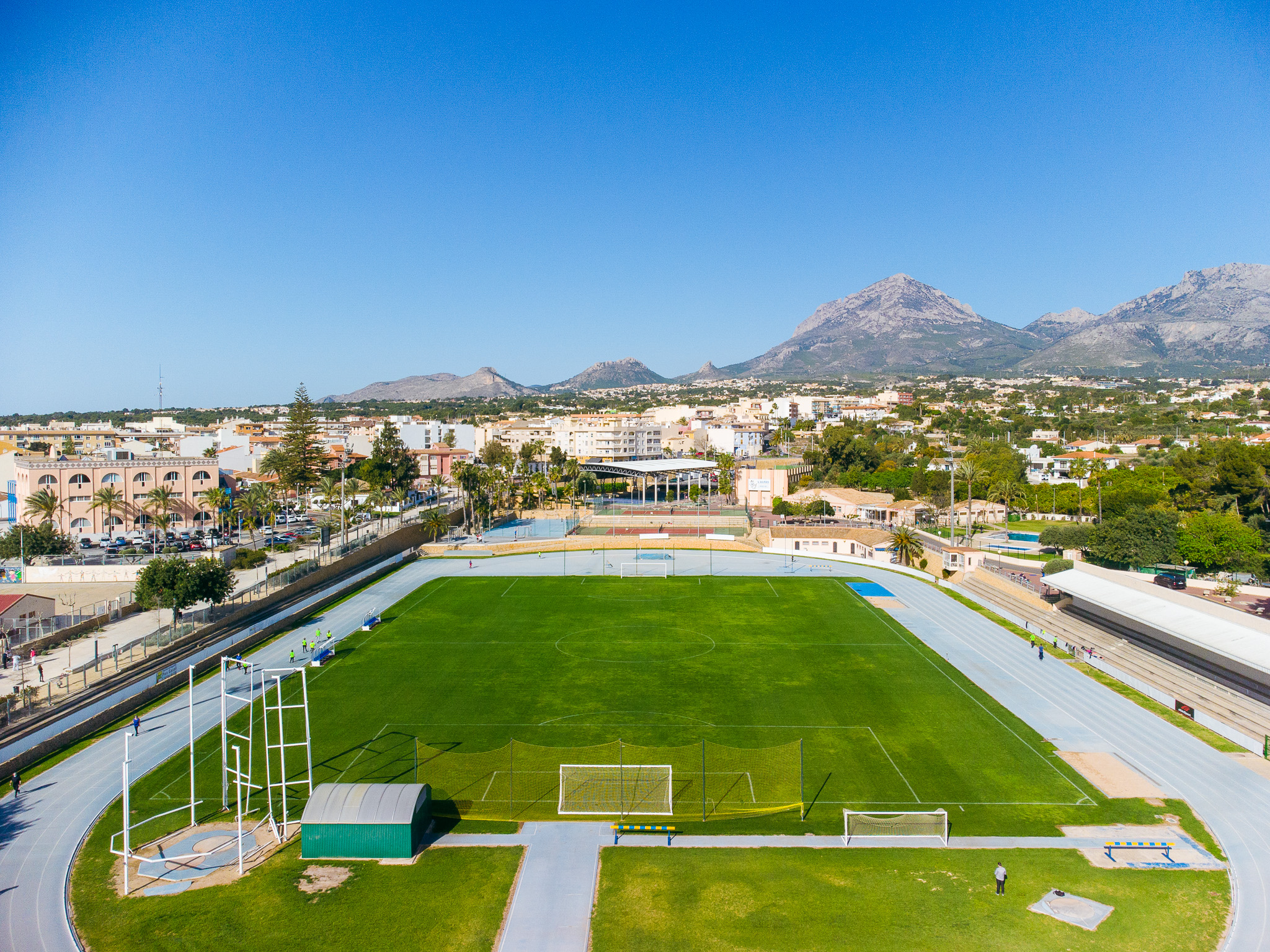 A well-kept stadium with a running track, a green pitch and a mountain panorama in the background.
