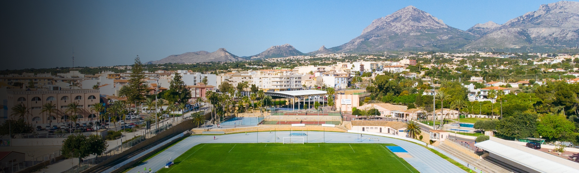 A well-kept stadium with a running track, a green pitch and a mountain panorama in the background.
