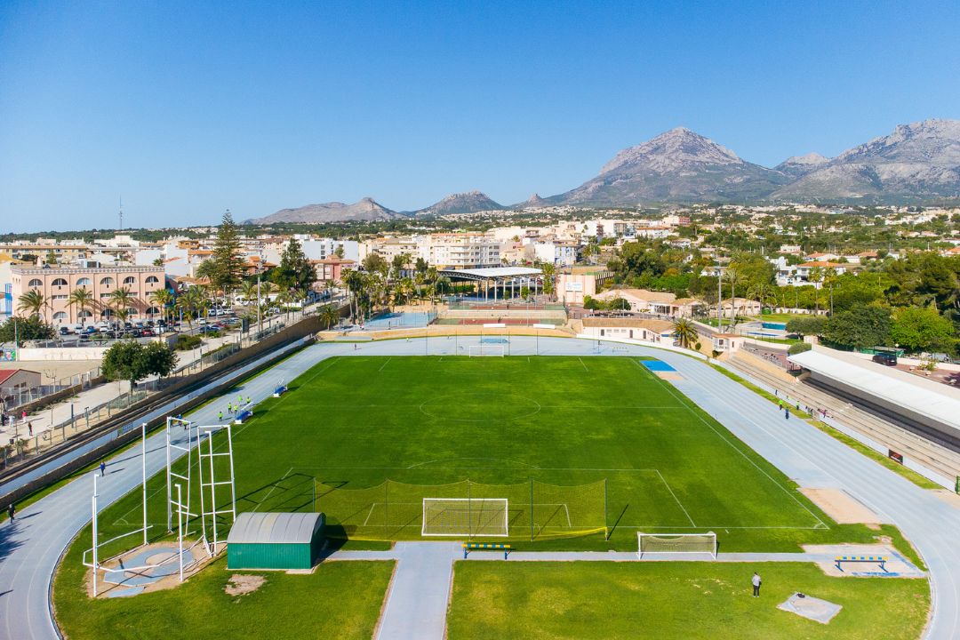 A well-kept stadium with a running track, a green pitch and a mountain panorama in the background.