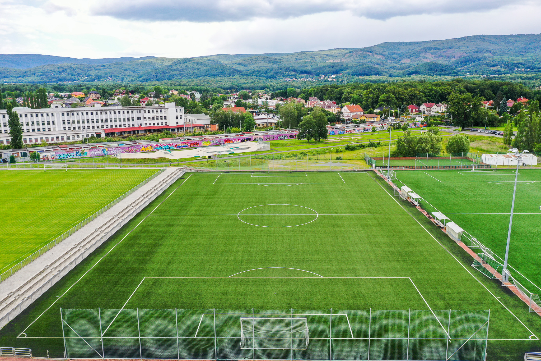 Fußballplatz mit Kunstrasen in einer grünen Umgebung, umgeben von Hügeln und einer Stadtlandschaft im Hintergrund.