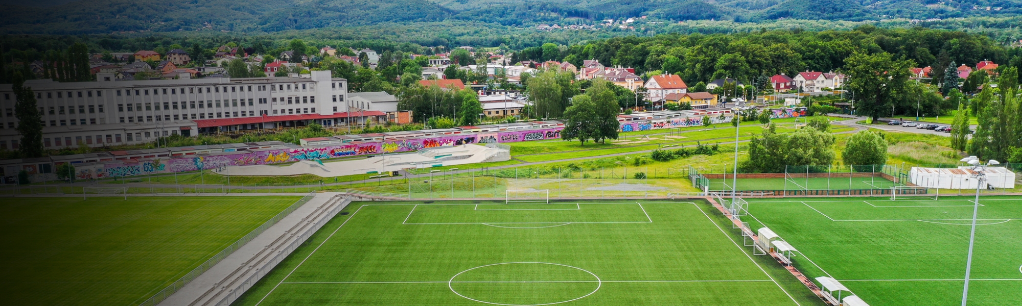 Fußballplatz mit Kunstrasen in einer grünen Umgebung, umgeben von Hügeln und einer Stadtlandschaft im Hintergrund.