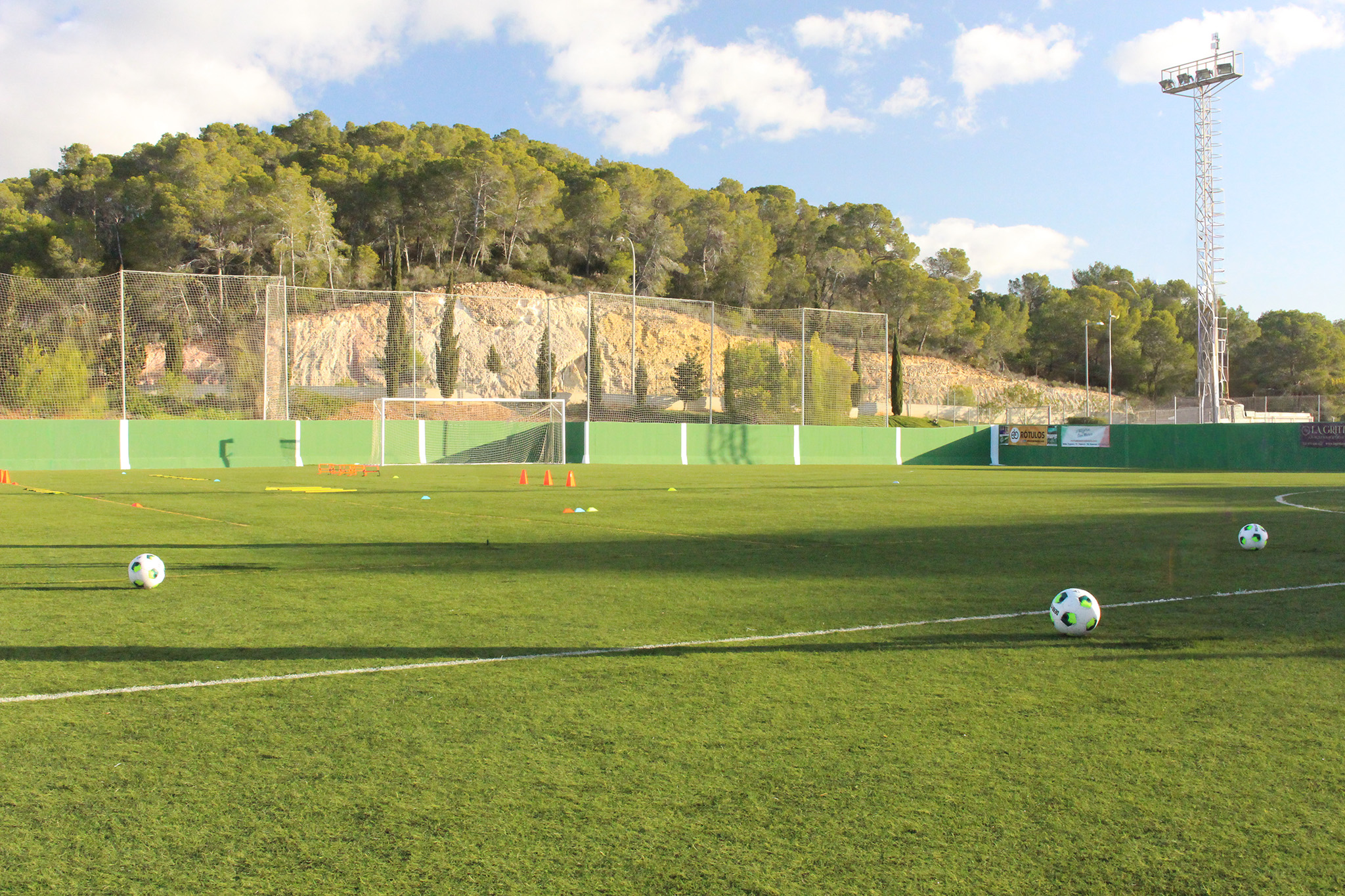 Fußballtrainingsplatz auf Sardinien mit grünem Kunstrasen, Tor, Bänken und Flutlichtmasten, umgeben von Netzen und Berglandschaft.