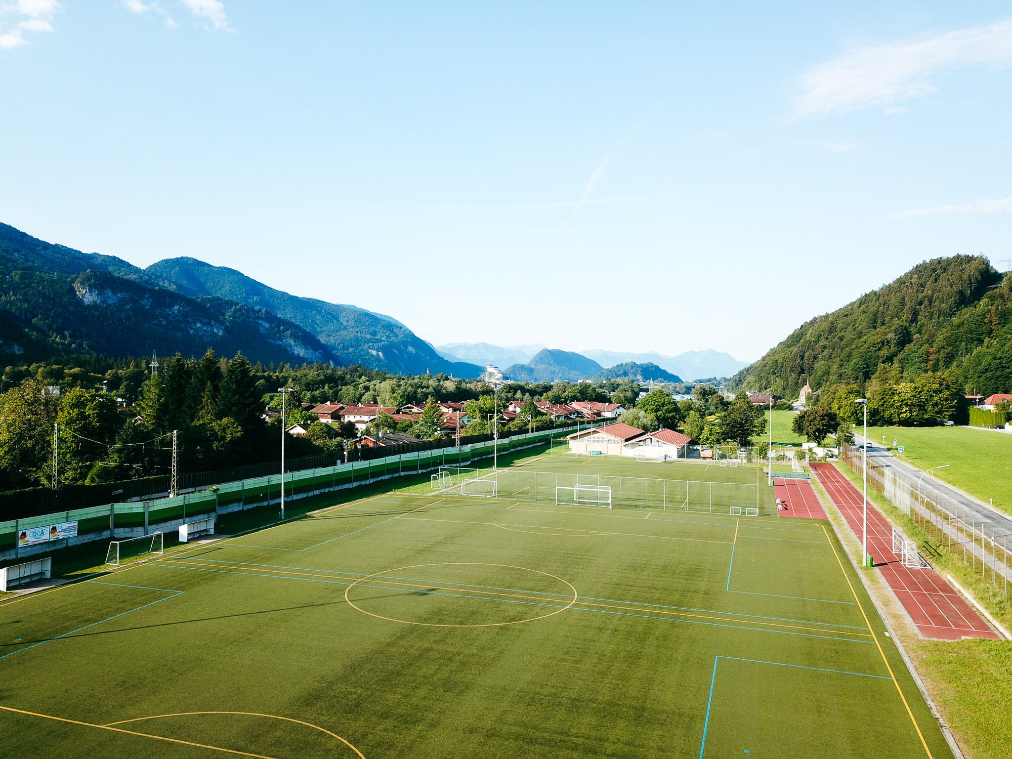 Kunstrasen-Fußballplatz mit einer kurzen Laufbahn daneben in Bayern, eingebettet in eine Alpenlandschaft mit malerischem Dorf im Hintergrund.