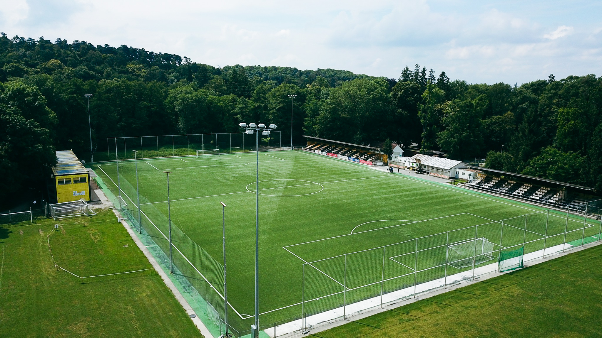 Fußballtrainingsplatz umgeben von dichten Wäldern mit einer kleinen Tribüne und Flutlichtmasten.