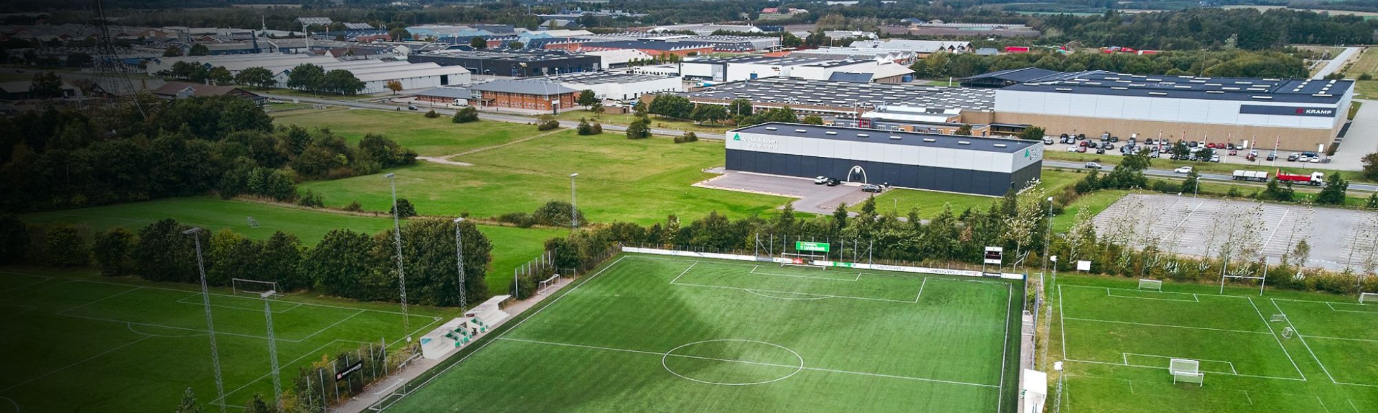 Panoramaaufnahme einer Fußballtrainingsanlage in Dänemark mit einem Kunstrasenplatz, mehreren Naturrasenplätzen, Flutlichtmasten und angrenzenden Industriehallen sowie Parkplätzen im Hintergrund.
