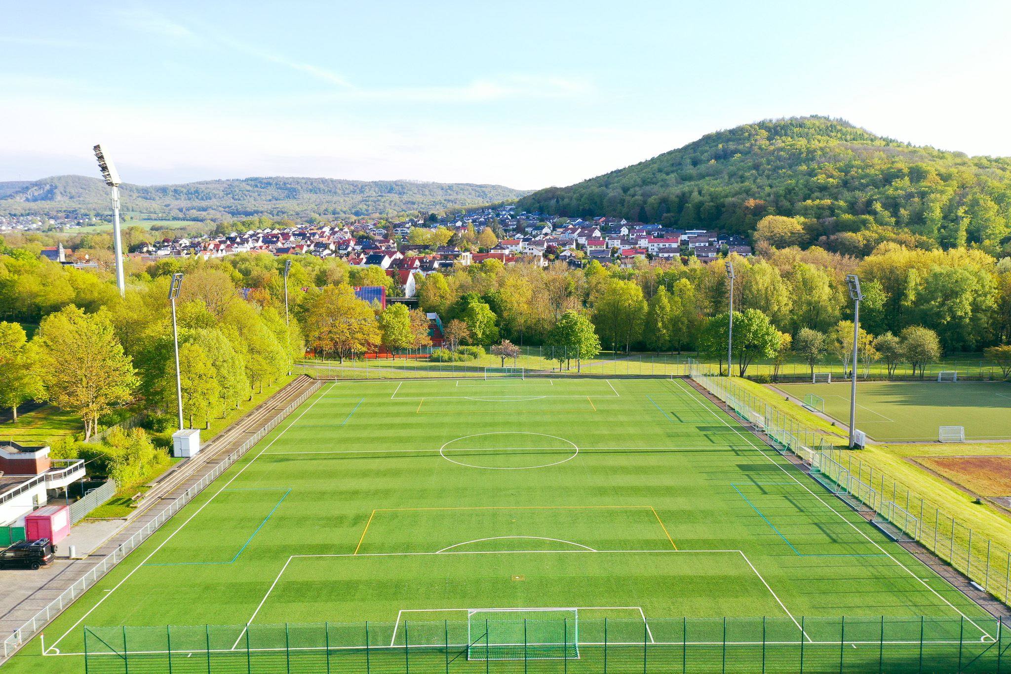 Luftaufnahme eines modernen Kunstrasen-Fußballplatzes mit Flutlichtanlage, umgeben von grünen Bäumen und Hügeln, im Hintergrund eine Kleinstadt mit roten Dächern in Hessen.