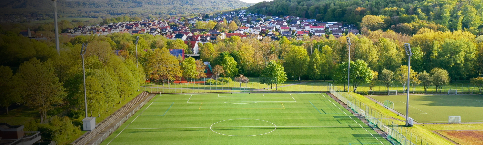 Fußballplatz mit Kunstrasen und Tribüne, eingebettet in eine grüne Landschaft mit Hügeln und einer Kleinstadt, optimal für ein Trainingslager in Hessen.