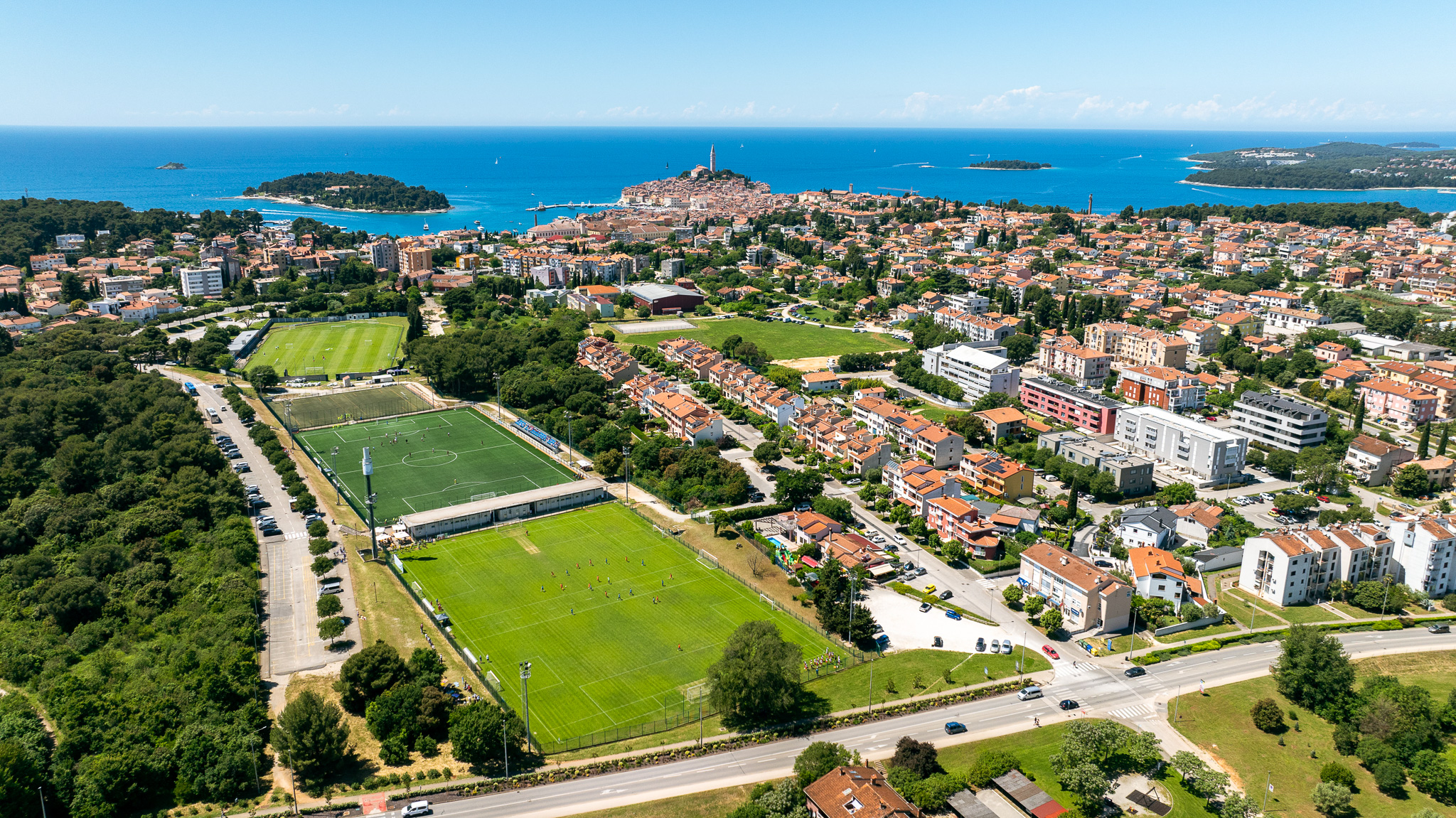 Aerial view of a football training facility in Croatia with several grass and artificial turf pitches, floodlight masts, surrounded by residential buildings and green areas, with a small town and the Adriatic Sea in the background.