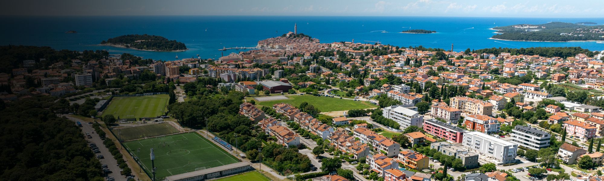 Panorma-Luftaufnahme einer Fußballtrainingsanlage in Kroatien mit mehreren Rasen- und Kunstrasenplätzen, Flutlichtmasten, umgeben von Wohnhäusern und Grünflächen, im Hintergrund eine kleine Stadt und die Adria.