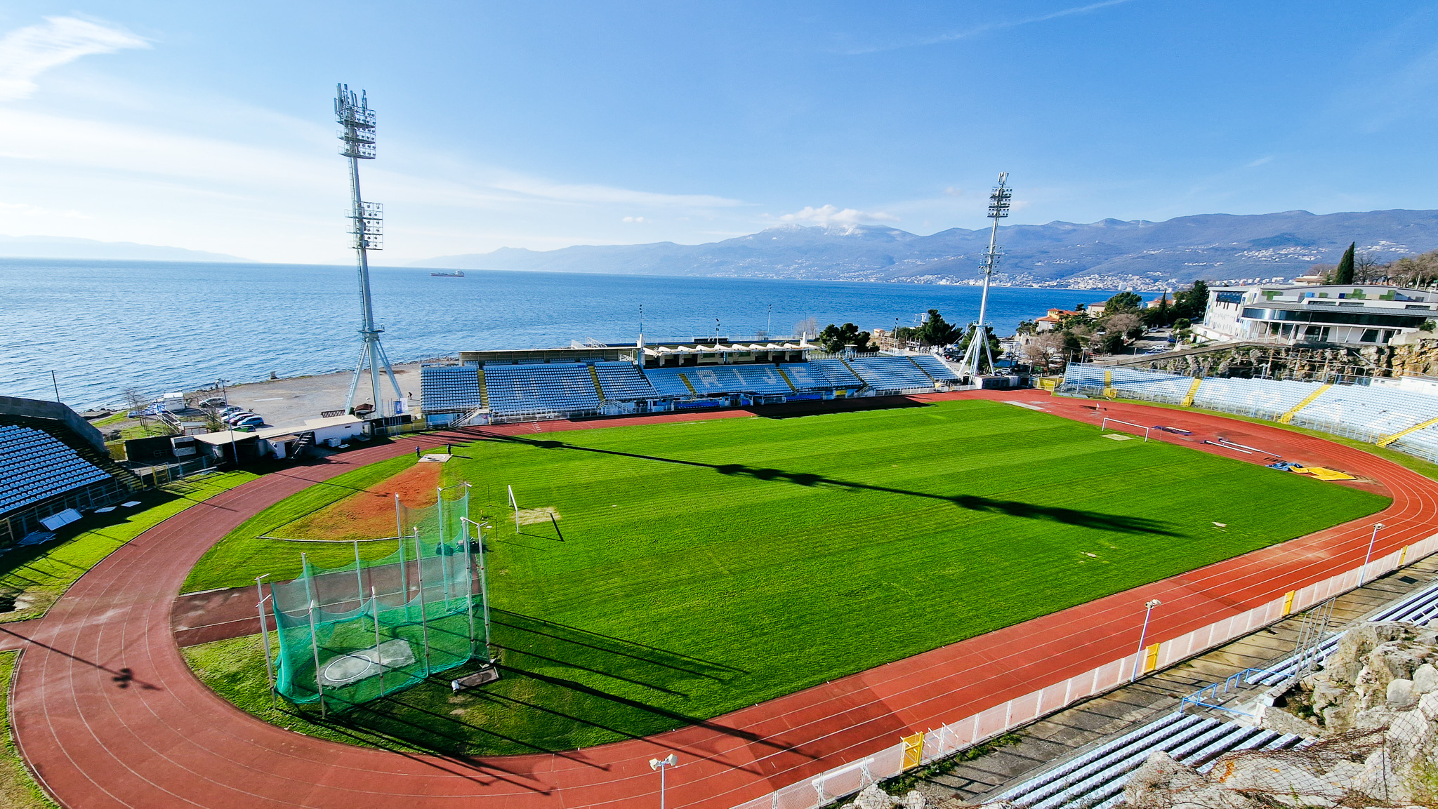 Aerial view of a football stadium on the Kvarner Bay in Croatia, located directly by the sea, with a running track and stands, surrounded by mountains and the Adriatic Sea in the background.