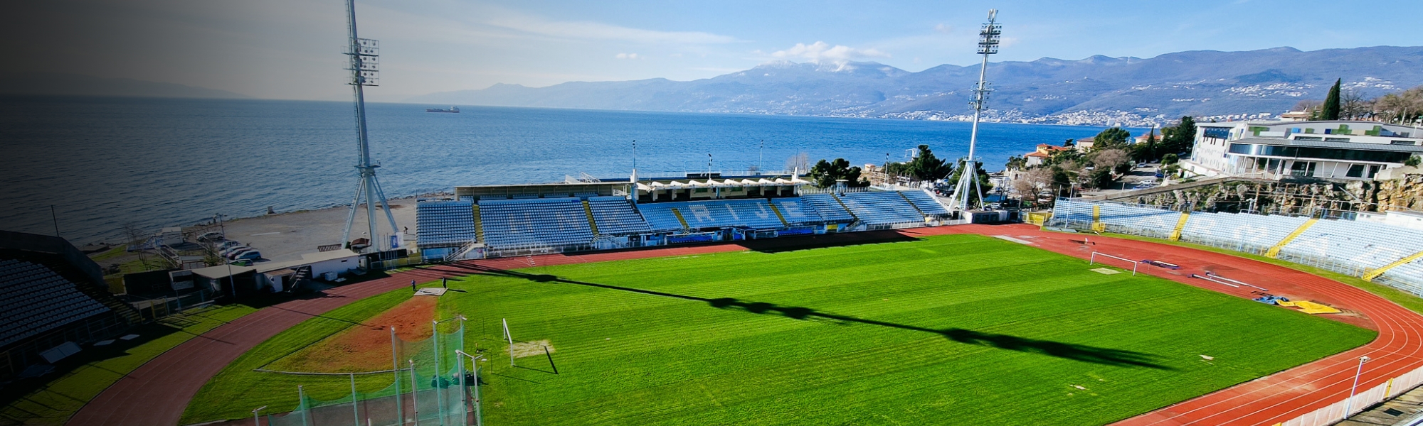 Panoramic view of a football stadium on Kvarner Bay, with a view of the pitch, the running track, the blue stands and the glittering sea, framed by a mountainous coastal landscape.