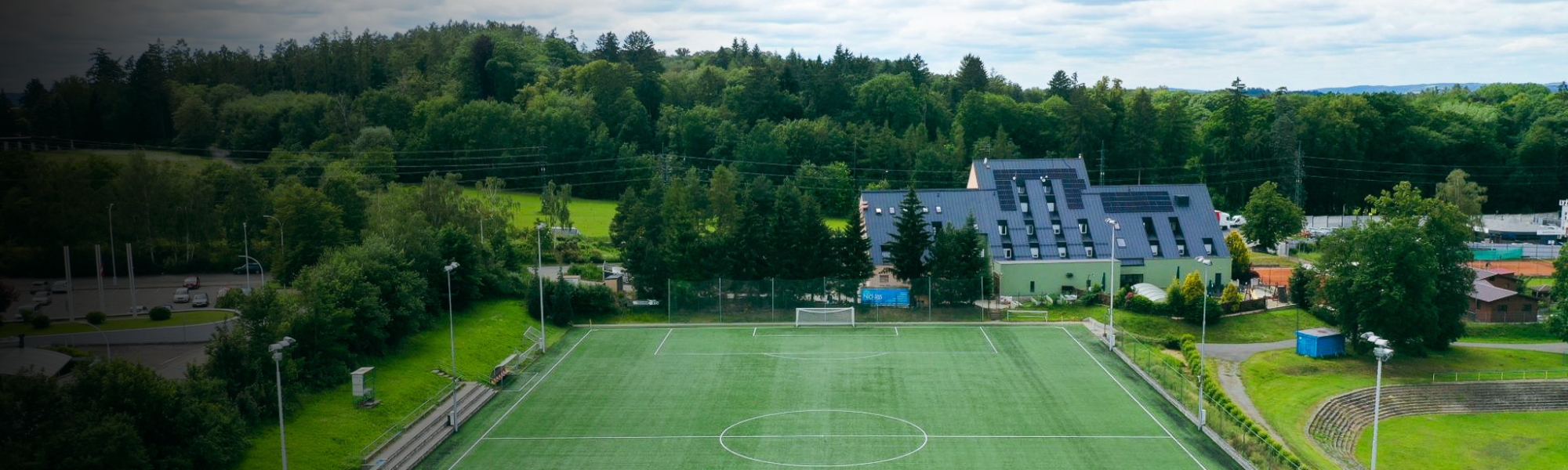 Fußballplatz mit Kunstrasen in einer idyllischen Umgebung, angrenzend an ein modernes Gebäude.