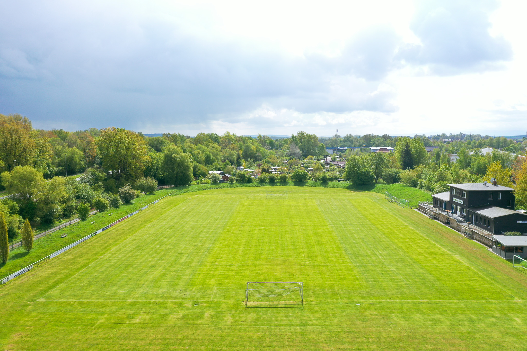Football pitch in Lower Saxony with freshly mown grass, surrounded by trees and a modern clubhouse with terrace.