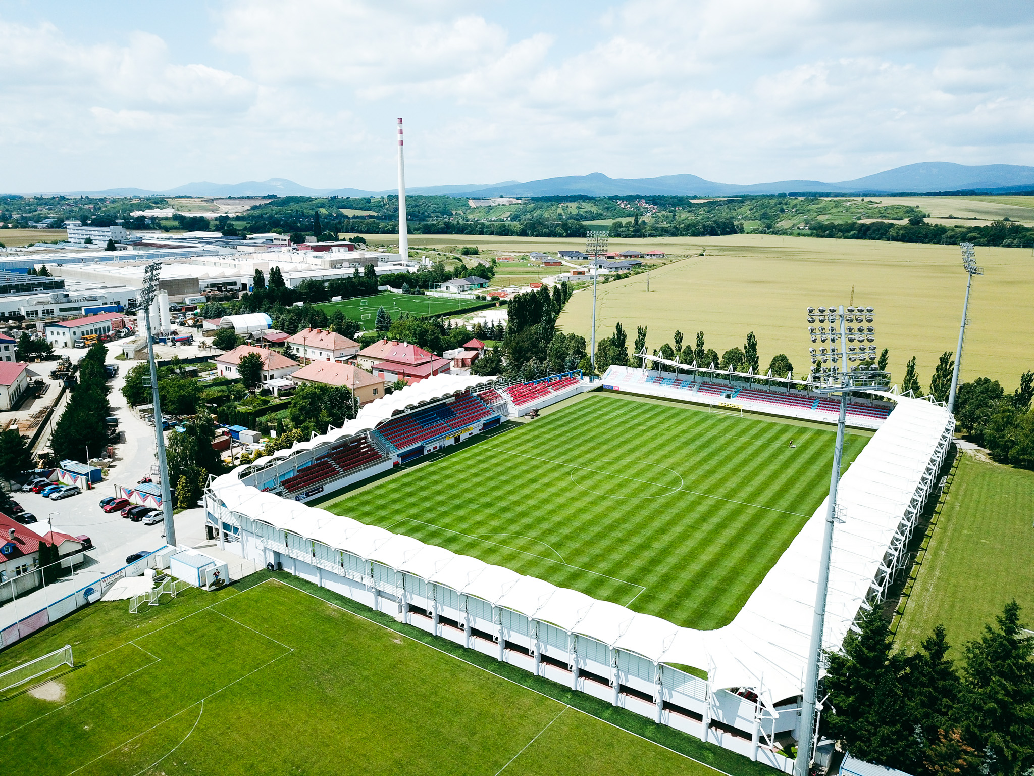 Fußballstadion in ländlicher Umgebung mit Tribünen, Flutlichtanlage und einer weiten Landschaft im Hintergrund.