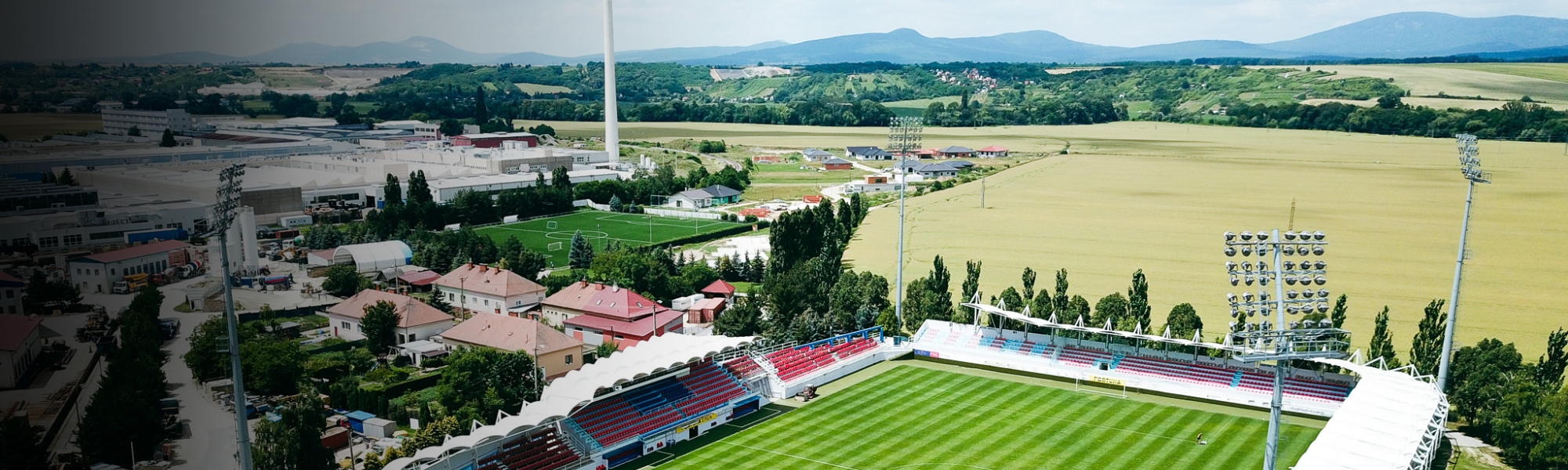 Fußballstadion in ländlicher Umgebung mit Tribünen, Flutlichtanlage und einer weiten Landschaft im Hintergrund.