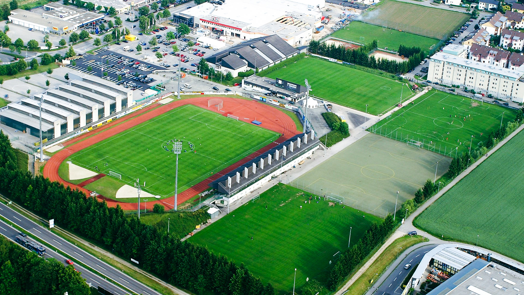Luftaufnahme einer modernen Sportanlage in Oberösterreich mit mehreren Fußballplätzen, darunter ein Stadion mit Laufbahn, Kunstrasen- und Rasenplätzen, umgeben von Parkplätzen, Gewerbebauten und Wohnhäusern.