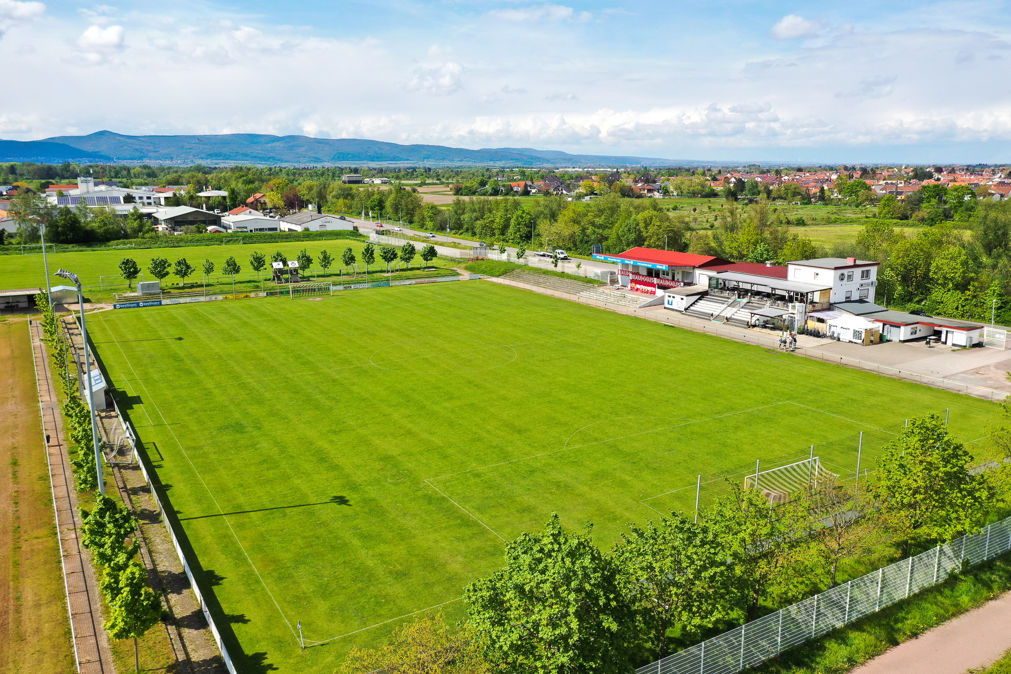Luftaufnahme eines gepflegten Naturrasen-Fußballplatzes mit angrenzendem Vereinsheim und Tribüne, eingebettet in eine grüne Landschaft mit Blick auf eine Stadt und Hügel im Hintergrund in Rheinland-Pfalz.