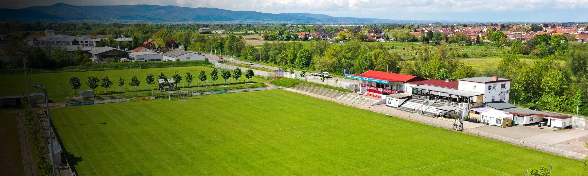 Weitläufiger Fußballplatz mit Naturrasen, Zuschauertribüne und Vereinsgebäude, umgeben von Bäumen und Feldern, mit Blick auf eine Kleinstadt in Rheinland-Pfalz.