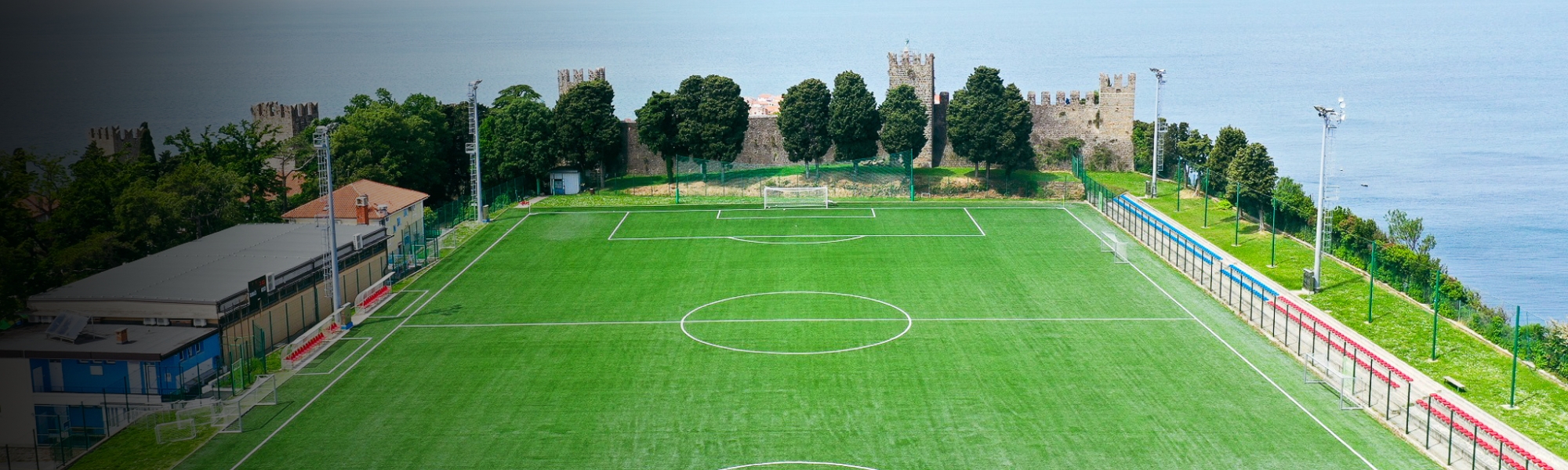 Luftaufnahme eines Fußballplatzes an der Küste, umgeben von grüner Landschaft und blauem Meer.