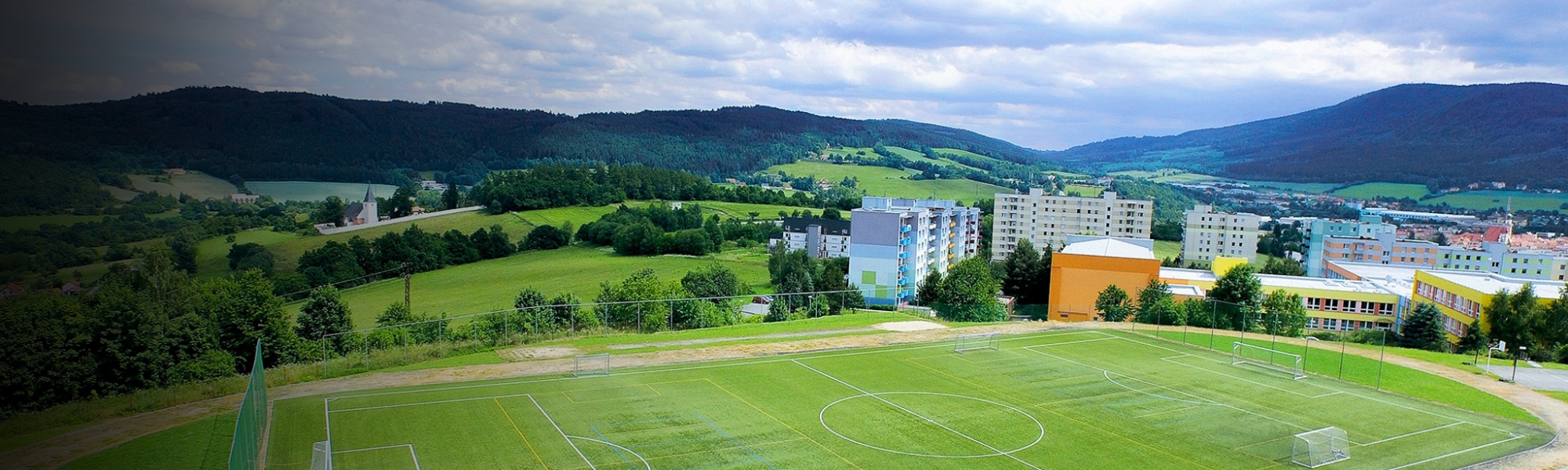 Fußballfeld mit Kunstrasen in einer malerischen Hügellandschaft, umgeben von Wohnhäusern.