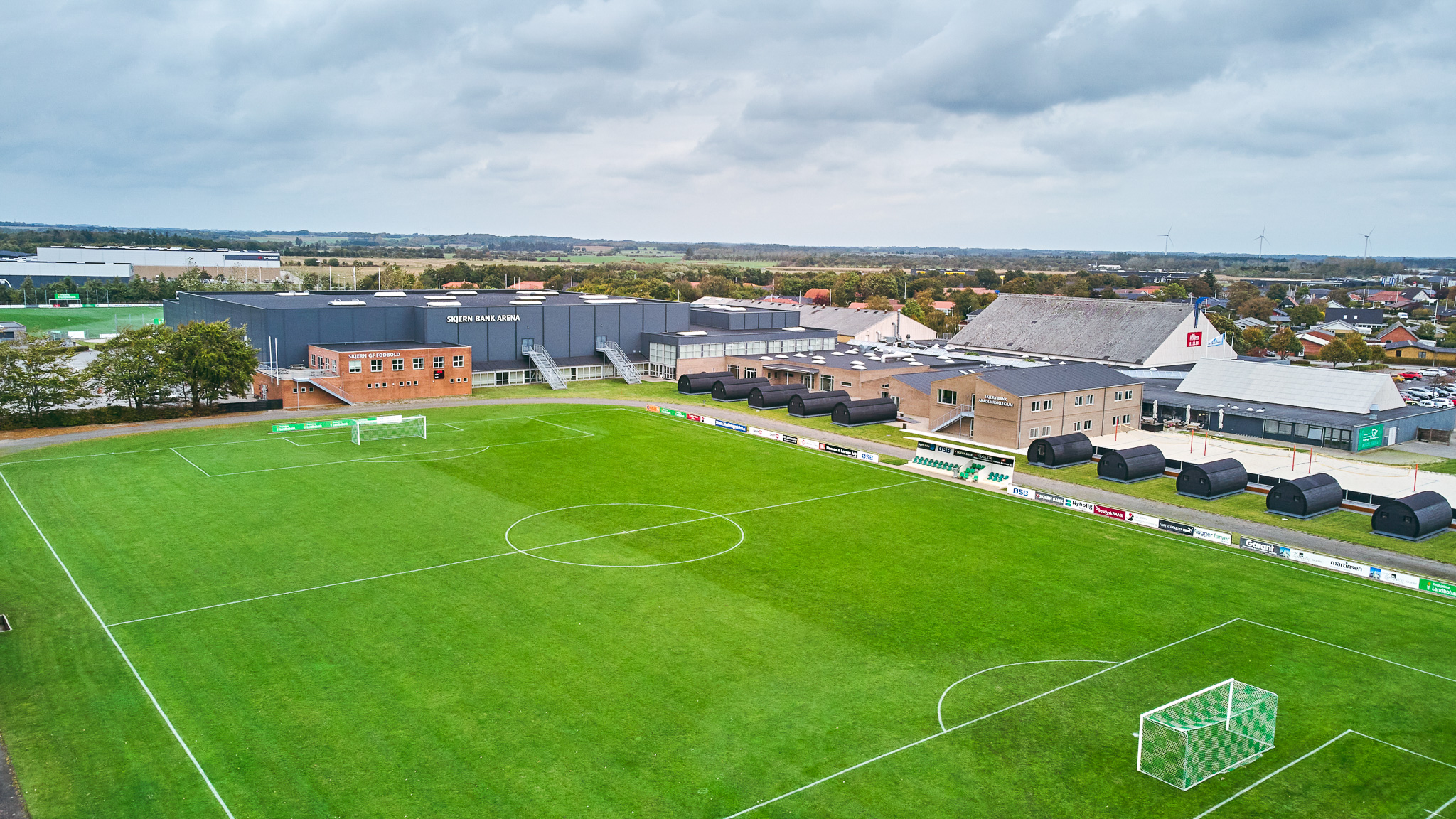 Fußballplatz mit angrenzender Sporthalle und modernen Unterkunftsmodulen in Süddänemark, umgeben von ländlicher Landschaft und kleinen Gebäuden.