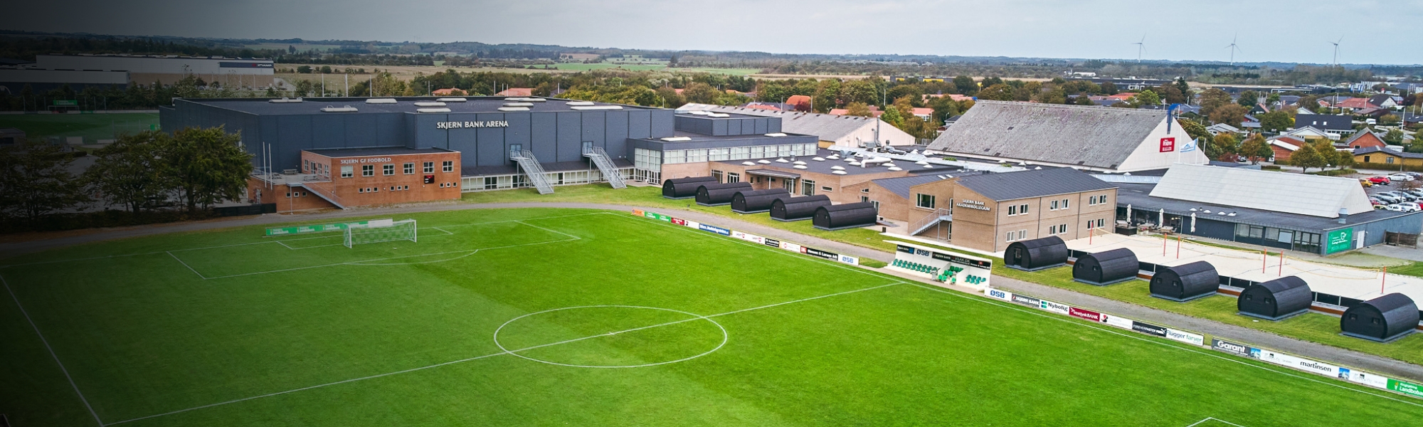 Sportanlage in Süddänemark mit Fußballplatz, Sitztribüne, Sporthalle und modernen Schlafmodulen, eingebettet in ein ländliches Umfeld mit Windrädern im Hintergrund.