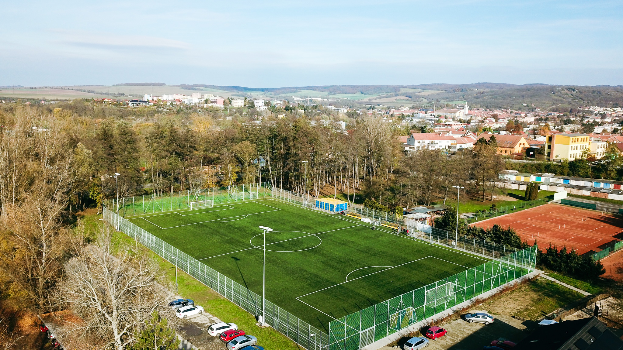 Fußballplatz mit Kunstrasen, gelegen in einer kleinen Stadt, umgeben von Wäldern und Tennisplätzen.